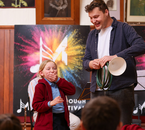 A little girl laughs at the sound a brass instrument made from a garden hose.